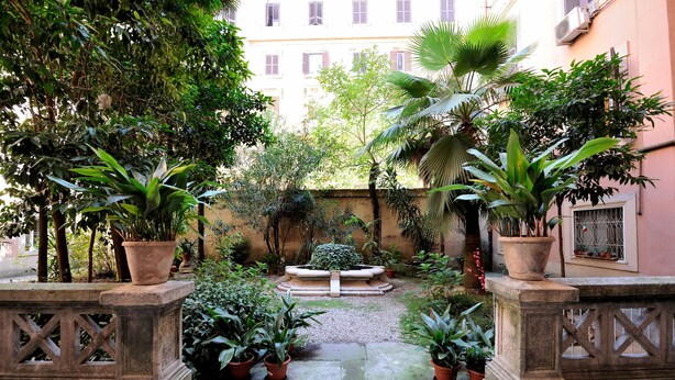 A courtyard with potted plants, a central fountain, and a building in the background. - White Vatican (Rome)