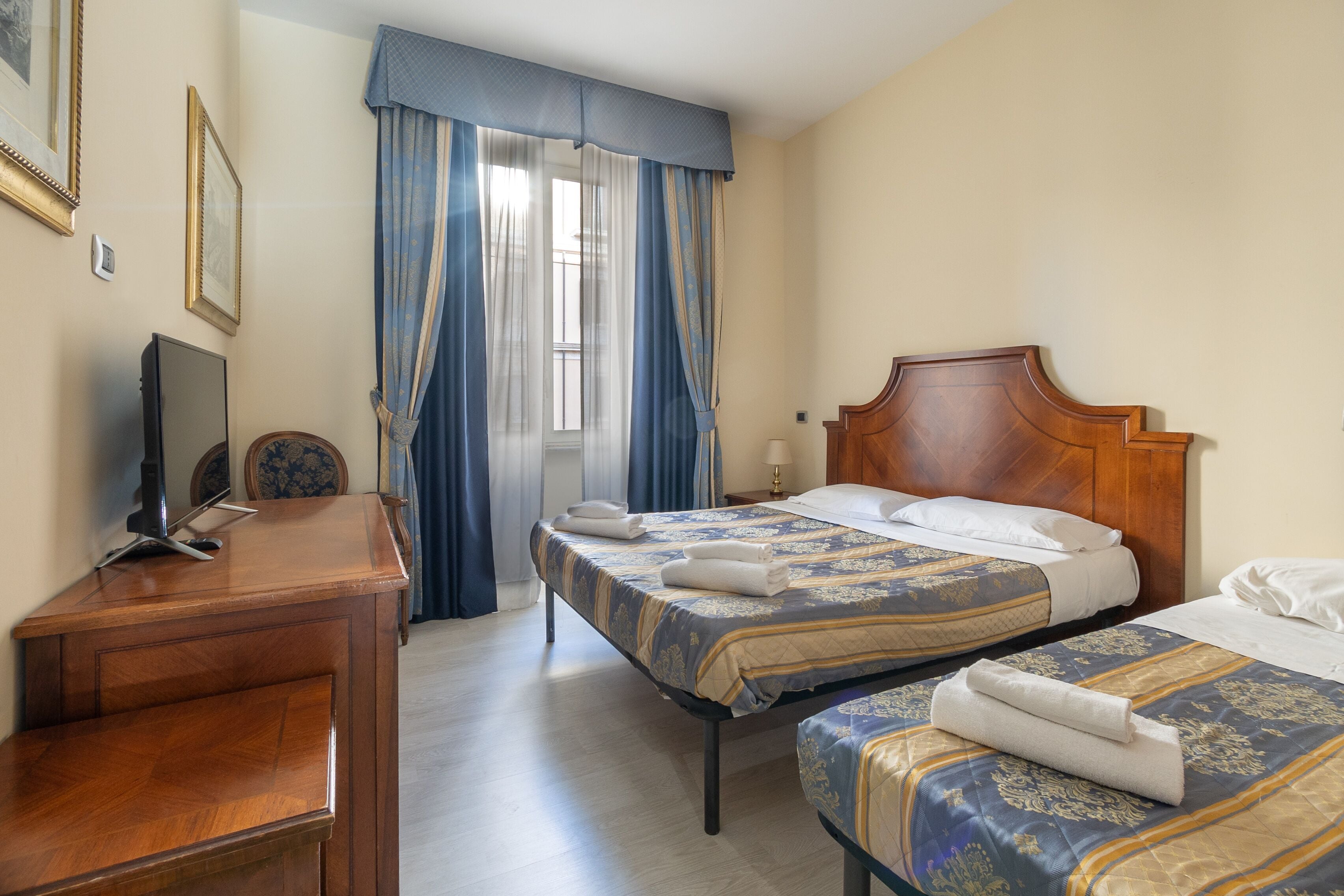 A hotel room with two beds, a desk, a TV, and a window with curtains.