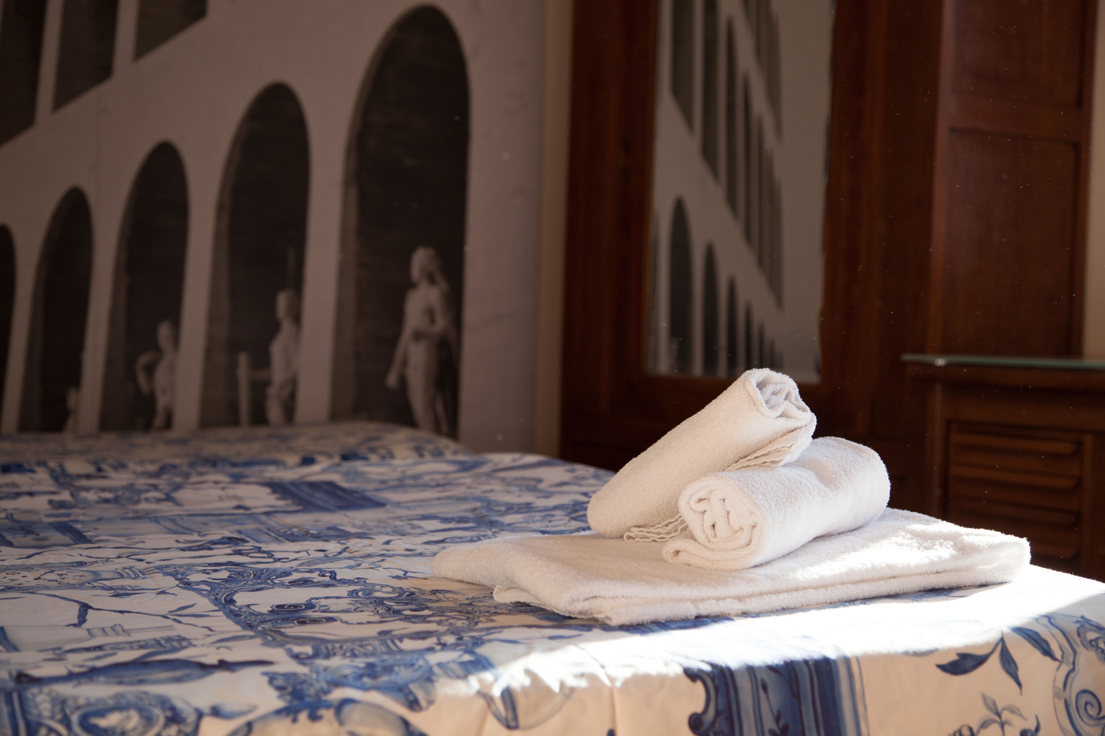 A bed with a blue and white patterned bedspread, a stack of neatly folded white towels, and a wall with a classical architectural mural.