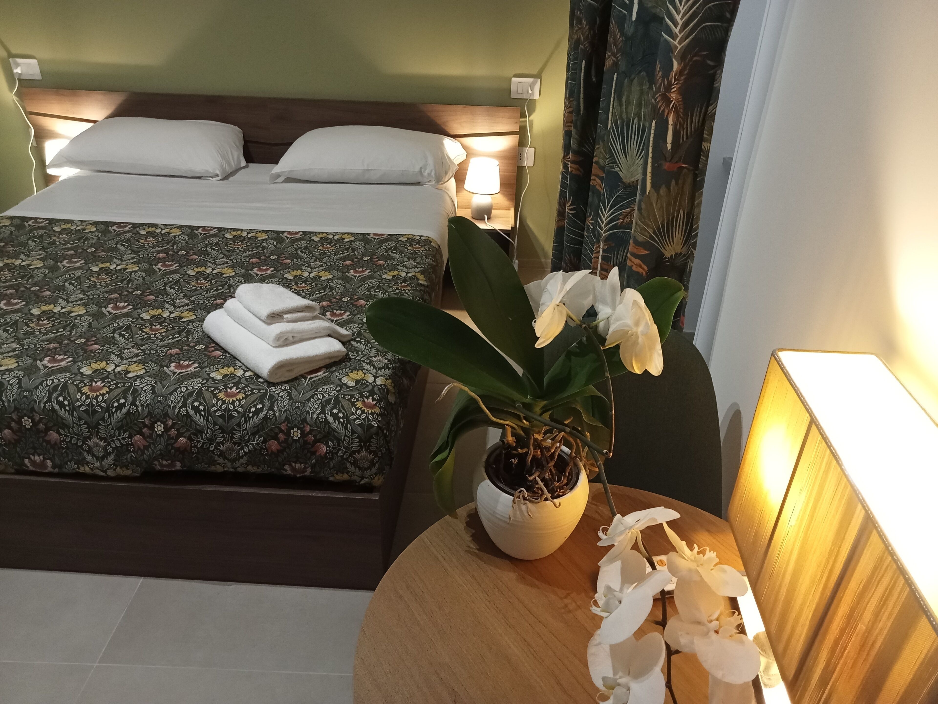 Un lit bien fait, avec des oreillers blancs et une couvre-lit à motifs floraux, une orchidée en pot sur une table de chevet et une table de nuit avec une lampe.