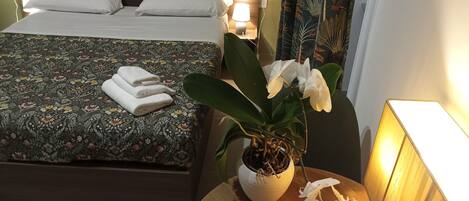 Un lit bien fait, avec des oreillers blancs et une couvre-lit à motifs floraux, une orchidée en pot sur une table de chevet et une table de nuit avec une lampe.