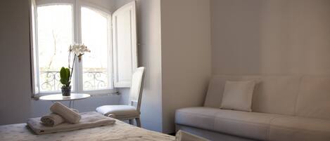 A hotel room with a bed, a sofa, a chair, a small table with a vase of flowers, and a window with a view of the outside.