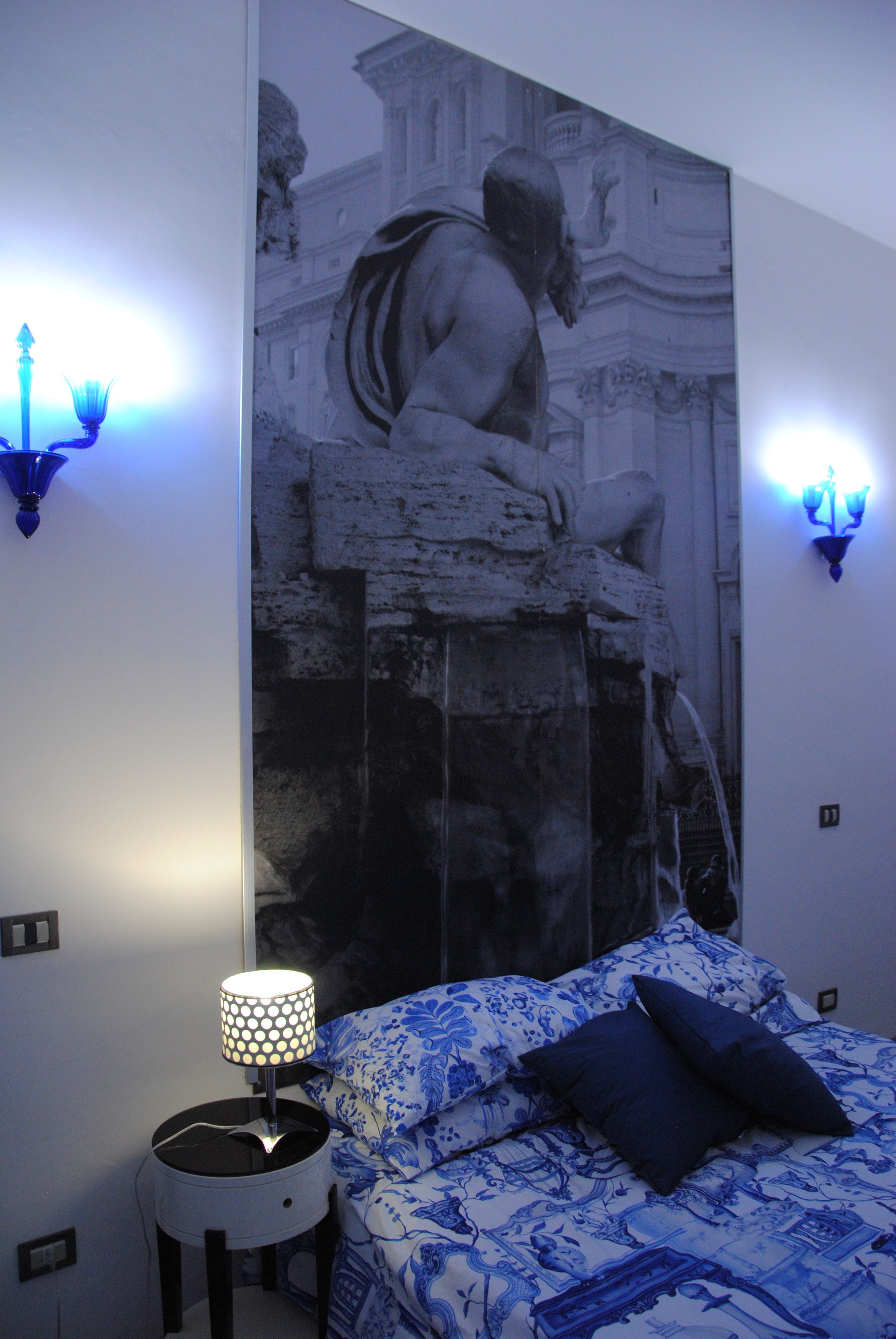 A bedroom with a bed, a nightstand, a wall-mounted lamp, and a large mural.
