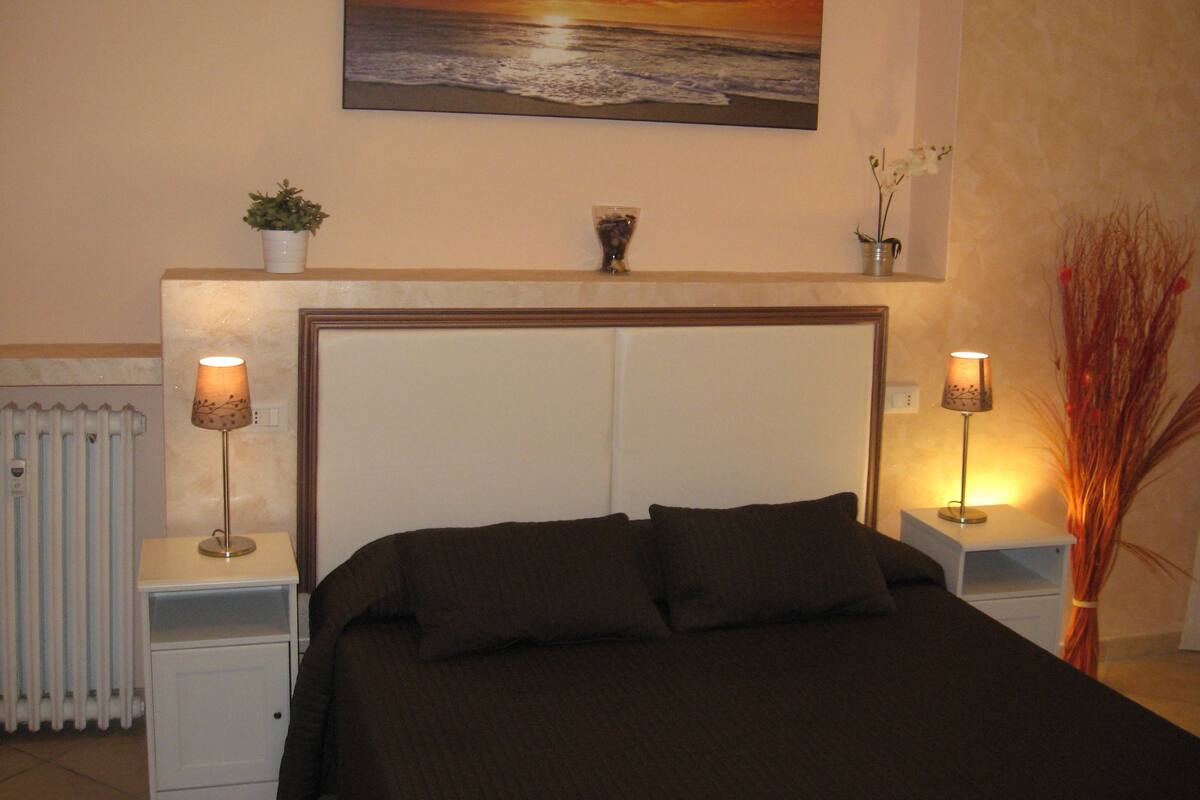 A bedroom with a large bed, two bedside tables with lamps, a radiator, and a wall-mounted painting of a sunset over the ocean.