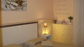 Un dormitorio con cama, mesita de noche, lámpara, decoración de pared y una pequeña estantería con una planta.
