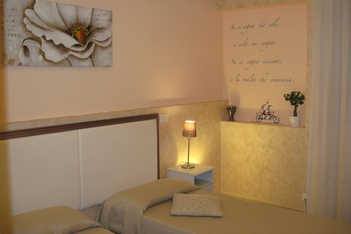 A bedroom with a bed, a nightstand, a lamp, a wall decoration, and a small shelf with a plant.