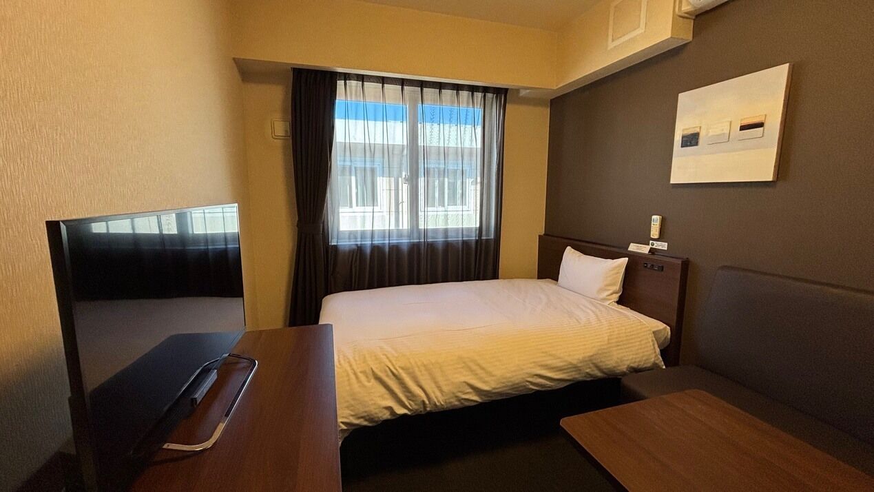 A hotel room with a bed, a desk, a chair, a television, and a window with curtains.