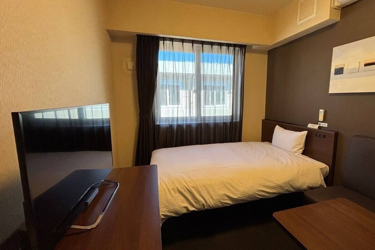 Habitación de hotel con cama, escritorio, silla, televisor y ventana con cortinas.