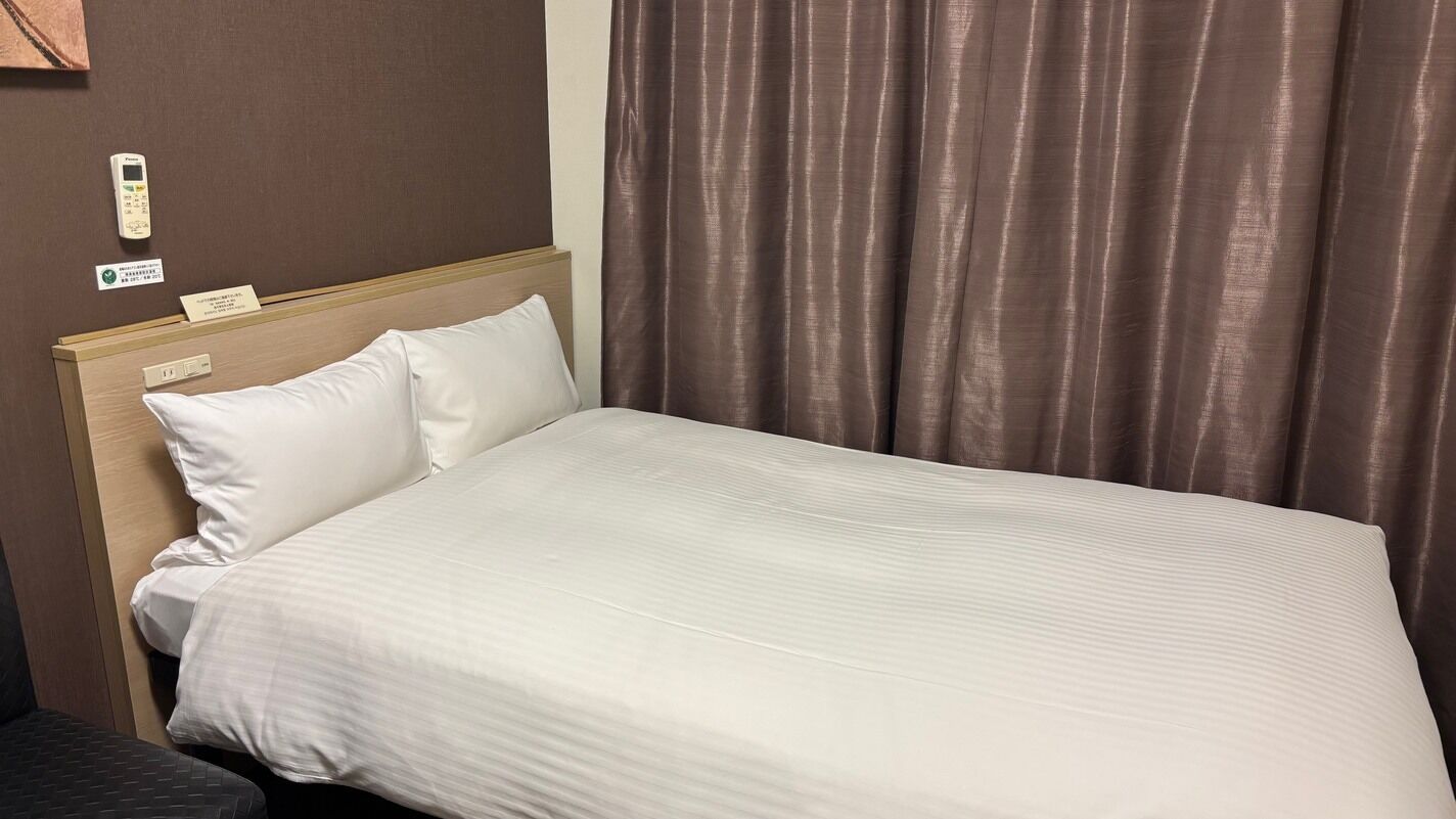 A hotel room with a bed, a bedside table, a control panel, and a curtain.