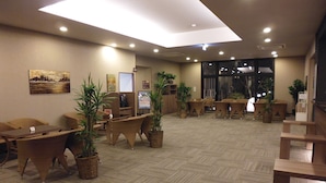 A hotel lobby with a herringbone-patterned floor, several wooden chairs and tables, potted plants, and a large window.