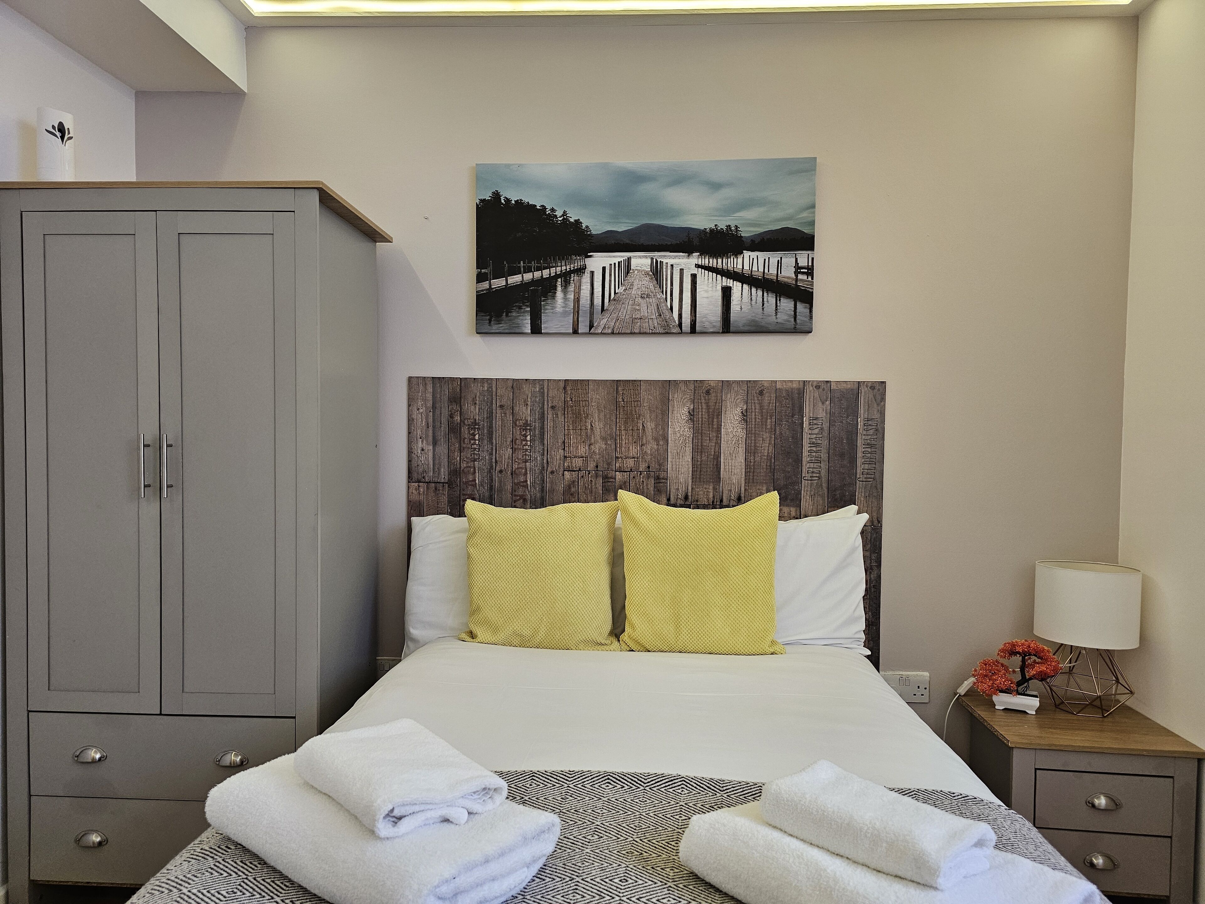 Un dormitorio ordenado con cama, cabecera de madera, armario gris y un cuadro con una escena de lago.