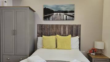 Un dormitorio ordenado con cama, cabecera de madera, armario gris y un cuadro con una escena de lago.