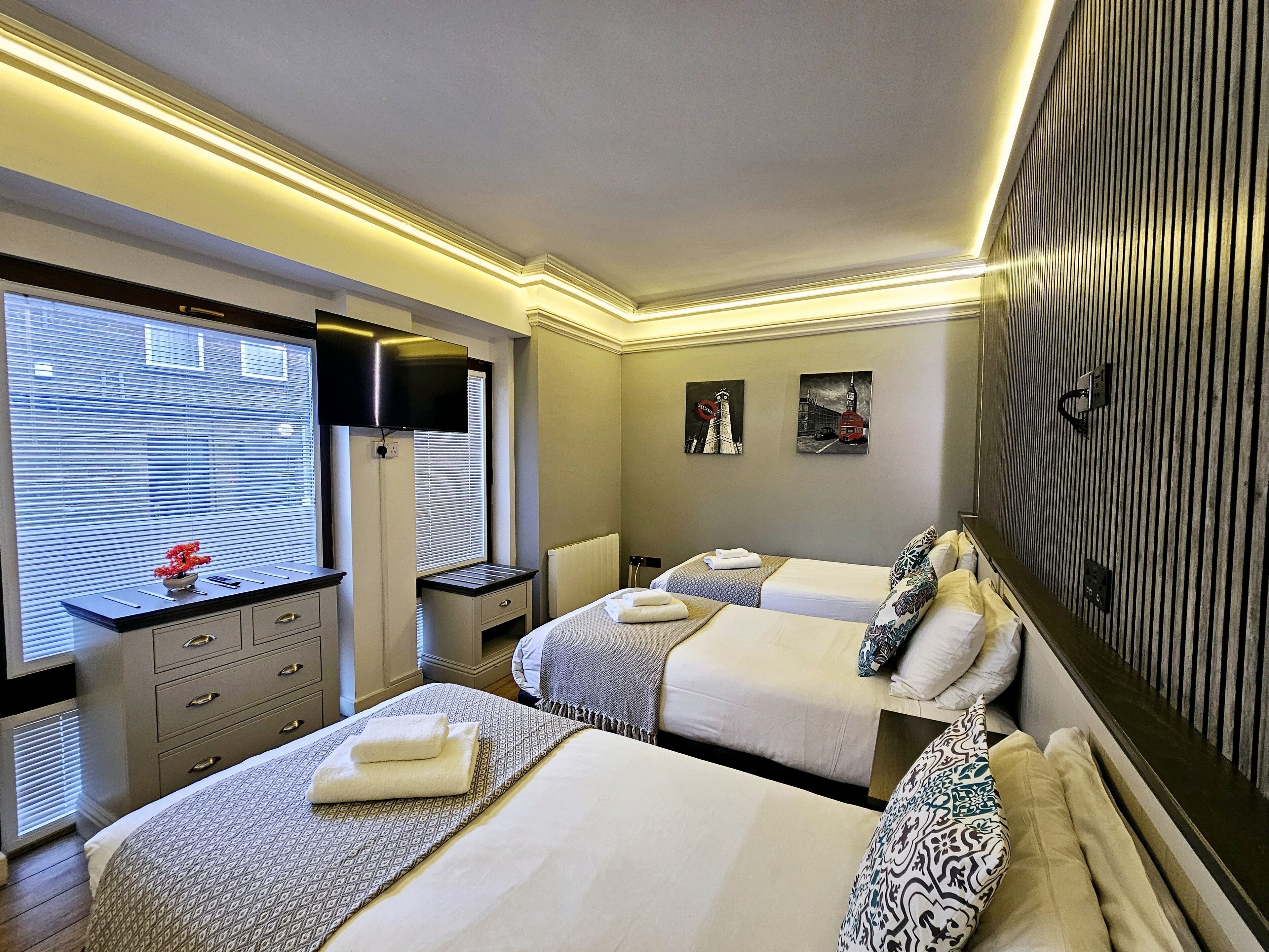 Habitación de hotel con dos camas, un escritorio y un televisor. Hay obras de arte en la pared y una ventana con persianas.