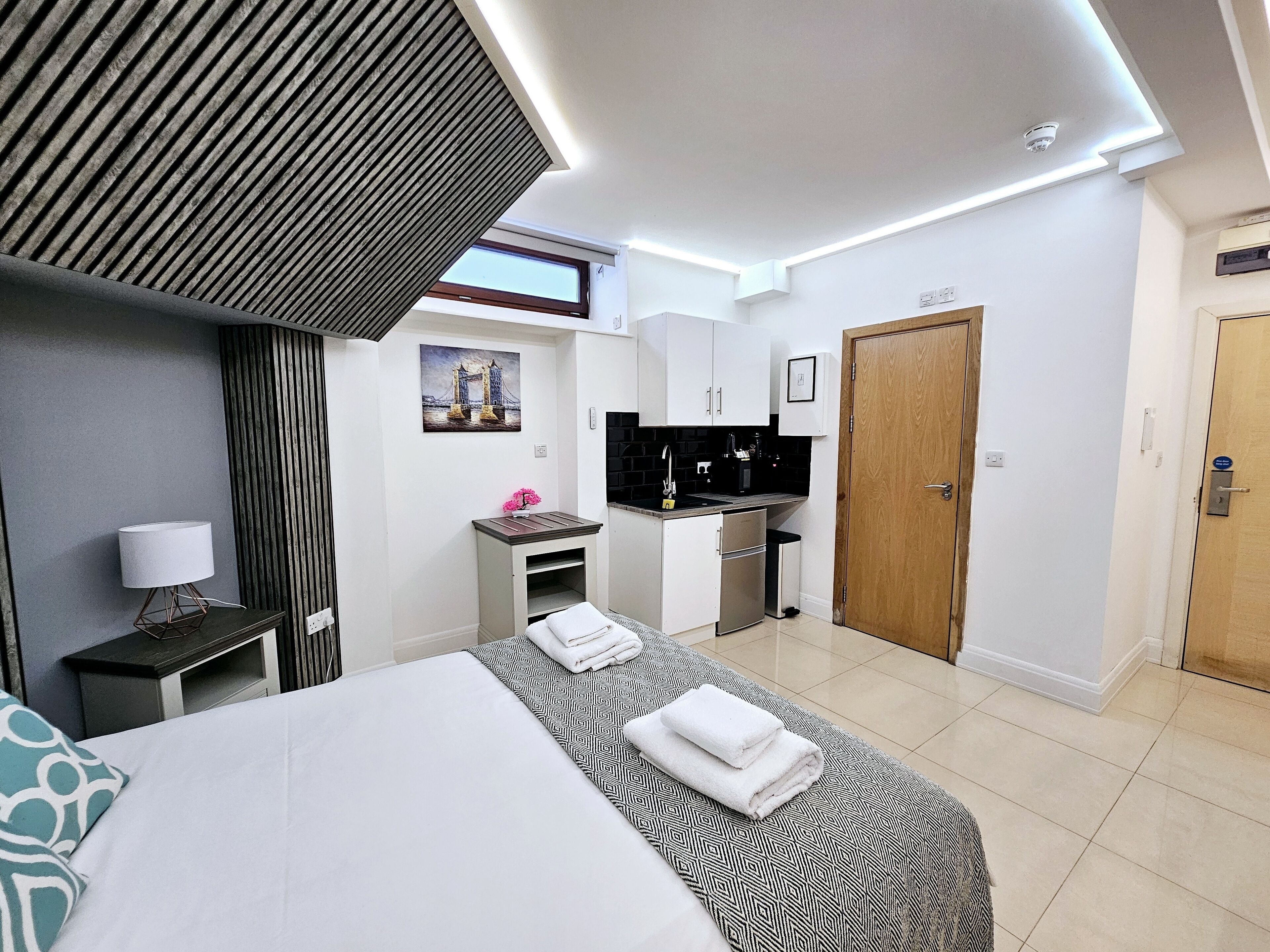 A hotel room with a bed, a desk with a lamp, a small kitchenette, a bathroom, and a door.