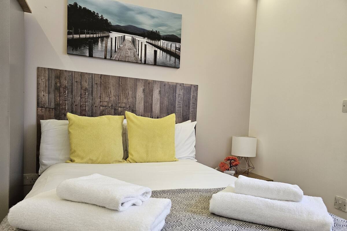 Ein Hotelzimmer mit einem hölzernen Kopfteil, einem Bett mit weißem und gelbem Kissen, einem gemusterten Teppich und einem Gemälde einer Brücke an der Wand.