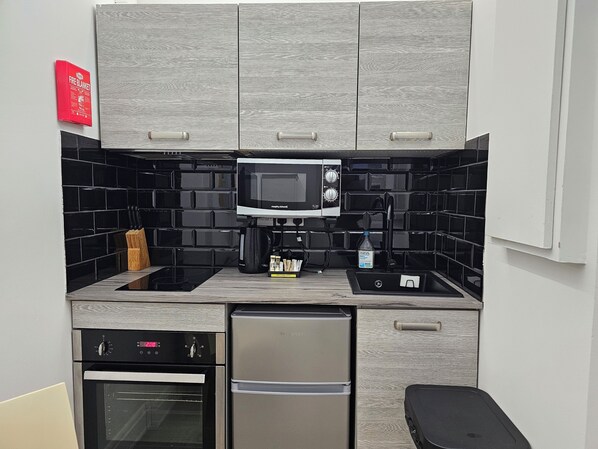 A compact kitchen with grey cabinets, a black tiled backsplash, a microwave, a coffee maker, a sink, a stove, and a refrigerator.