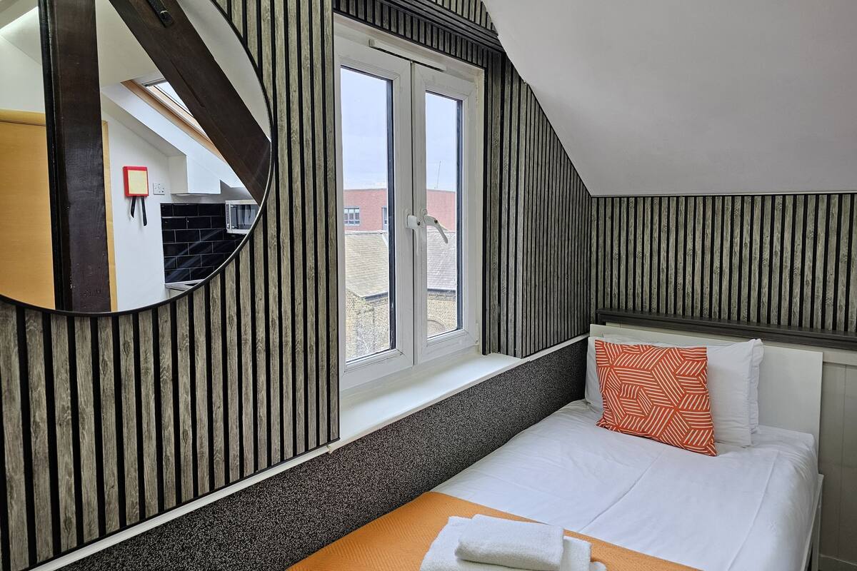 Ein Schlafzimmer mit großem Fenster, einem Bett mit weißen und orangefarbenen Kissen, einem runden Spiegel und einer Holzwandverkleidung.
