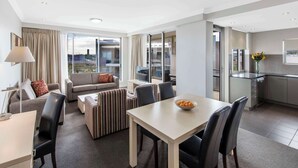 A modern hotel room with a dining area, a sofa set, a small kitchen, and large windows offering a city view.