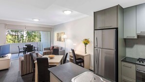 A modern kitchen with a stainless steel refrigerator, a dining area with a table and chairs, and a large window providing a view of greenery outside.