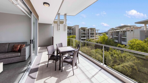 A balcony with a sofa and table, glass railings, and a view of other buildings and greenery.