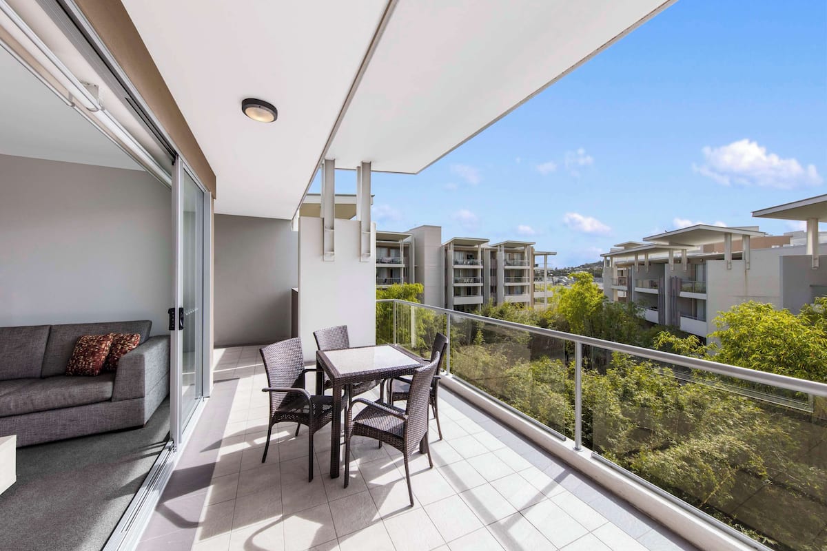 A balcony with a sofa and table, glass railings, and a view of other buildings and greenery.