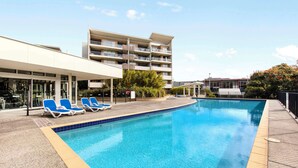 A swimming pool with lounge chairs, a building with multiple balconies, and a clear sky.