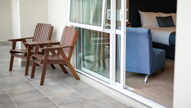 A balcony with two wooden chairs and a blue sofa, a sliding glass door, and a bed with a dark headboard and patterned pillows. - The Riverstone (Rangiora)