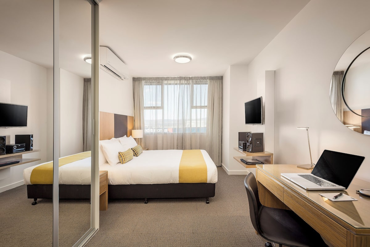 Quarto de hotel com cama, escrivaninha, laptop, televisão e uma janela grande com cortinas.
