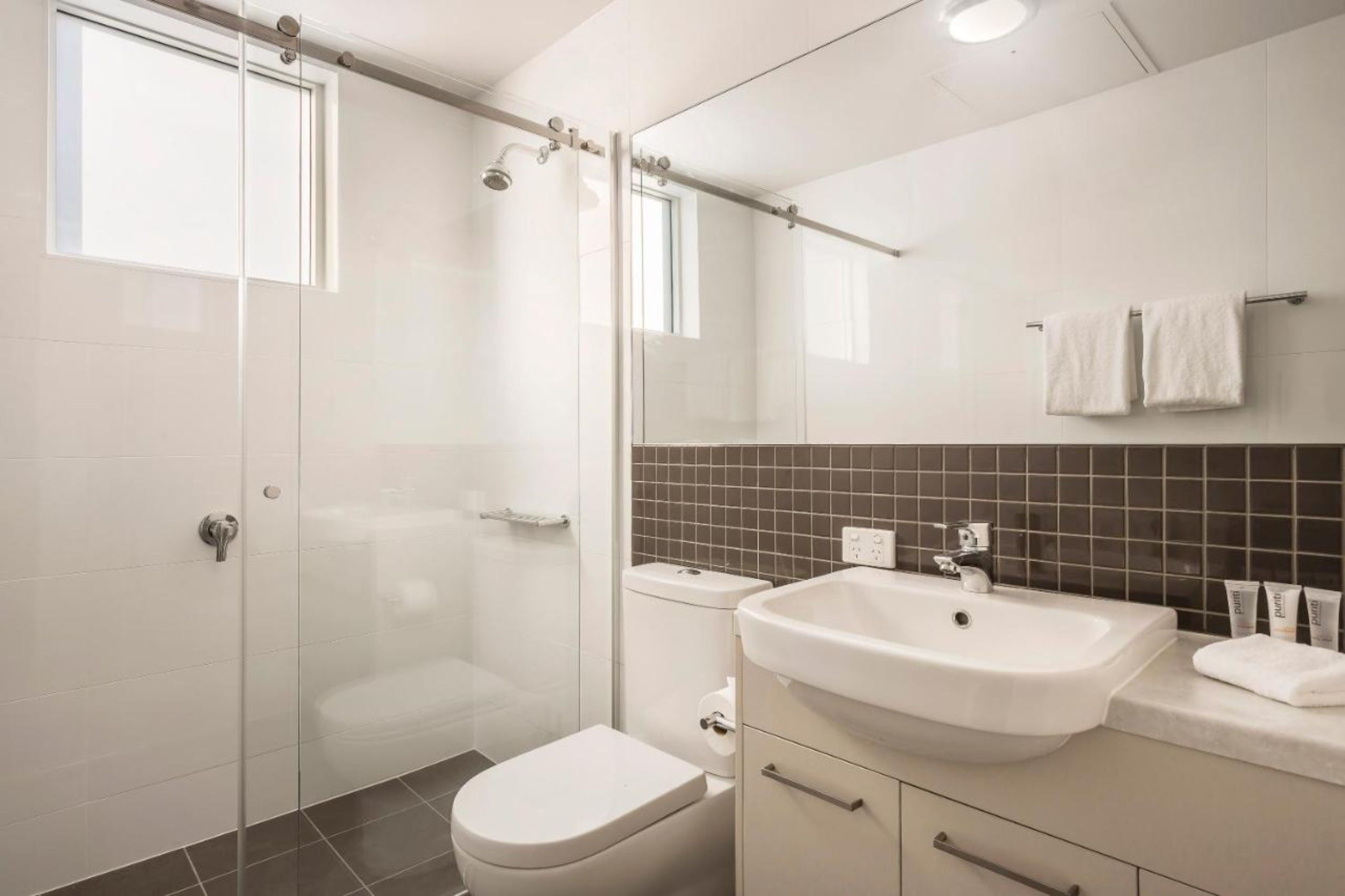 Une salle de bain moderne avec une douche à parois de verre, des toilettes blanches, un lavabo avec un miroir et un mur recouvert de carreaux.