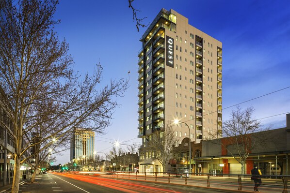 A multi-story hotel with a modern design, featuring balconies on each floor and a prominent "QUEST" sign on the facade.