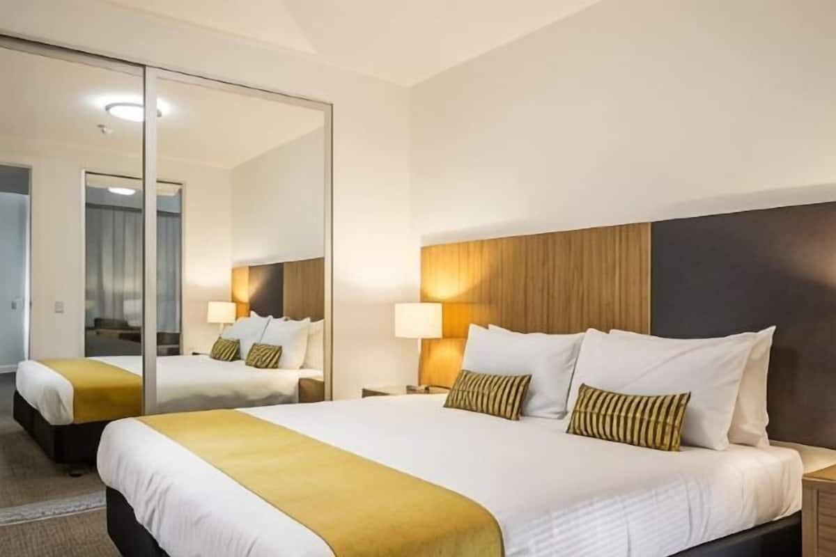 Quarto de hotel com duas camas, um espelho grande, abajures e cabeceira de madeira.