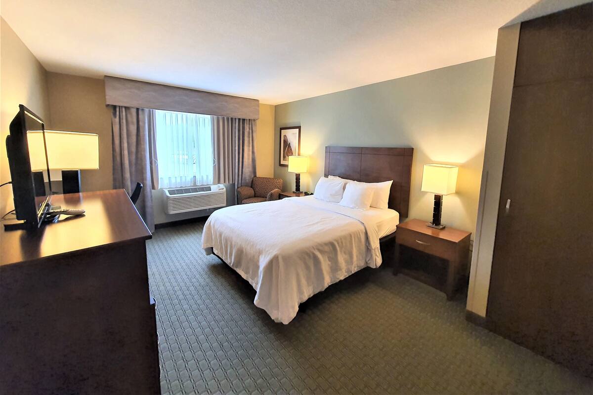 A hotel room with a bed, a desk, a chair, a window with curtains, and a ceiling fan.