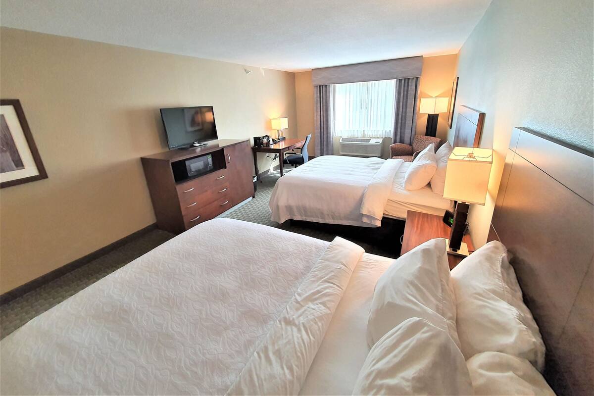 A hotel room with two beds, a TV on a dresser, a desk with a lamp, and a window with curtains.