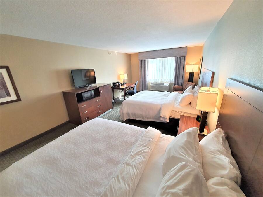 A hotel room with two beds, a TV on a dresser, a desk with a lamp, and a window with curtains.
