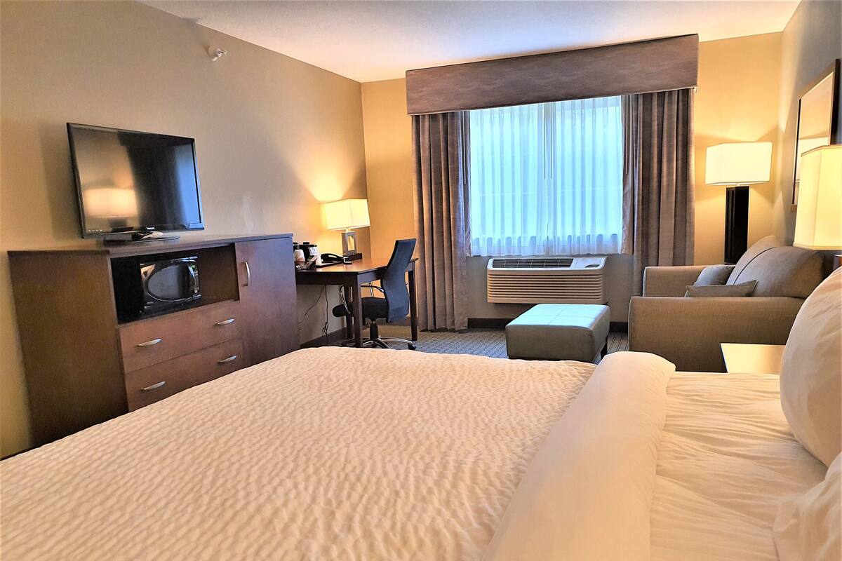 A hotel room with a large bed, a desk with a chair, a television, an air conditioning unit, and a window with curtains.