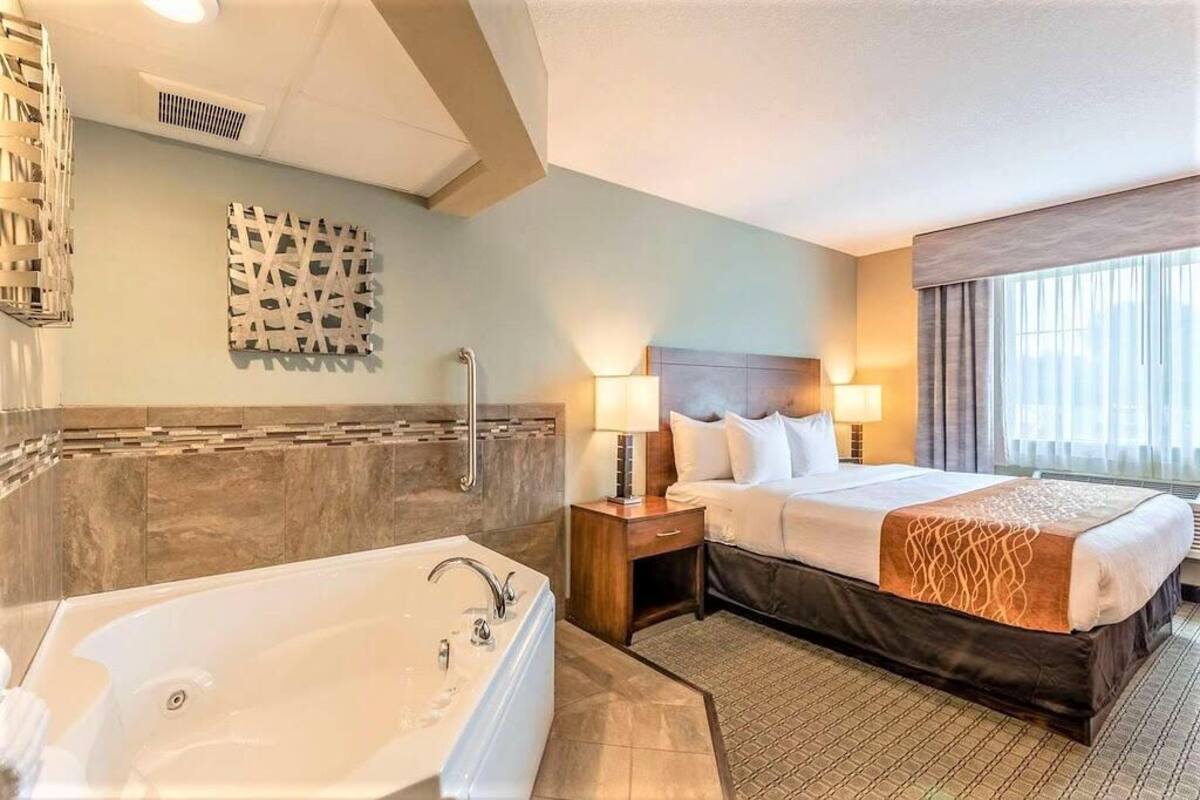 A hotel room with a large bathtub, a bed with two pillows, a nightstand with a lamp, and a window with curtains.