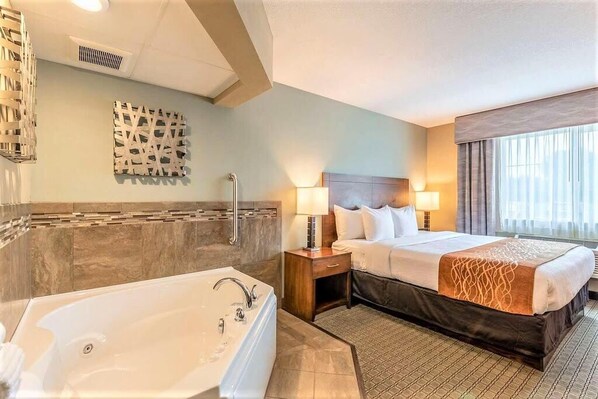 Habitación de hotel con una bañera grande, cama con dos almohadas, mesita de noche con lámpara y ventana con cortinas.
