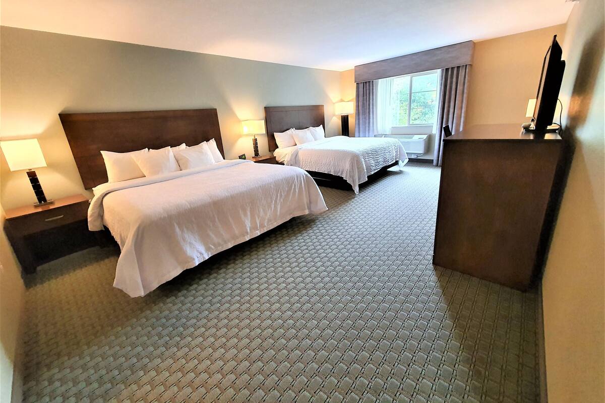 A hotel room with two beds, a large window, and a flat-screen TV on a stand.