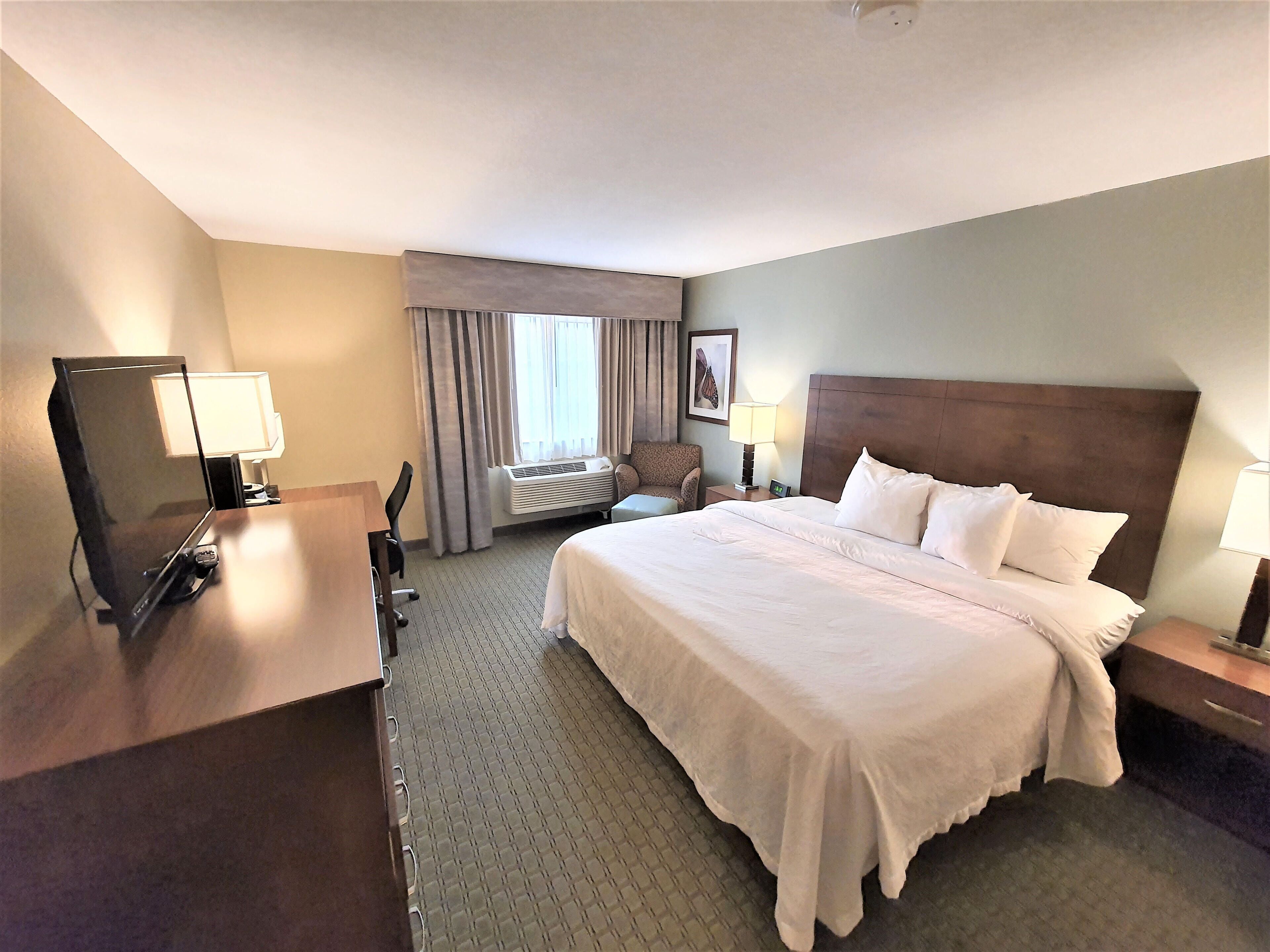 A hotel room with a large bed, a desk with a computer, a chair, and a window with curtains.