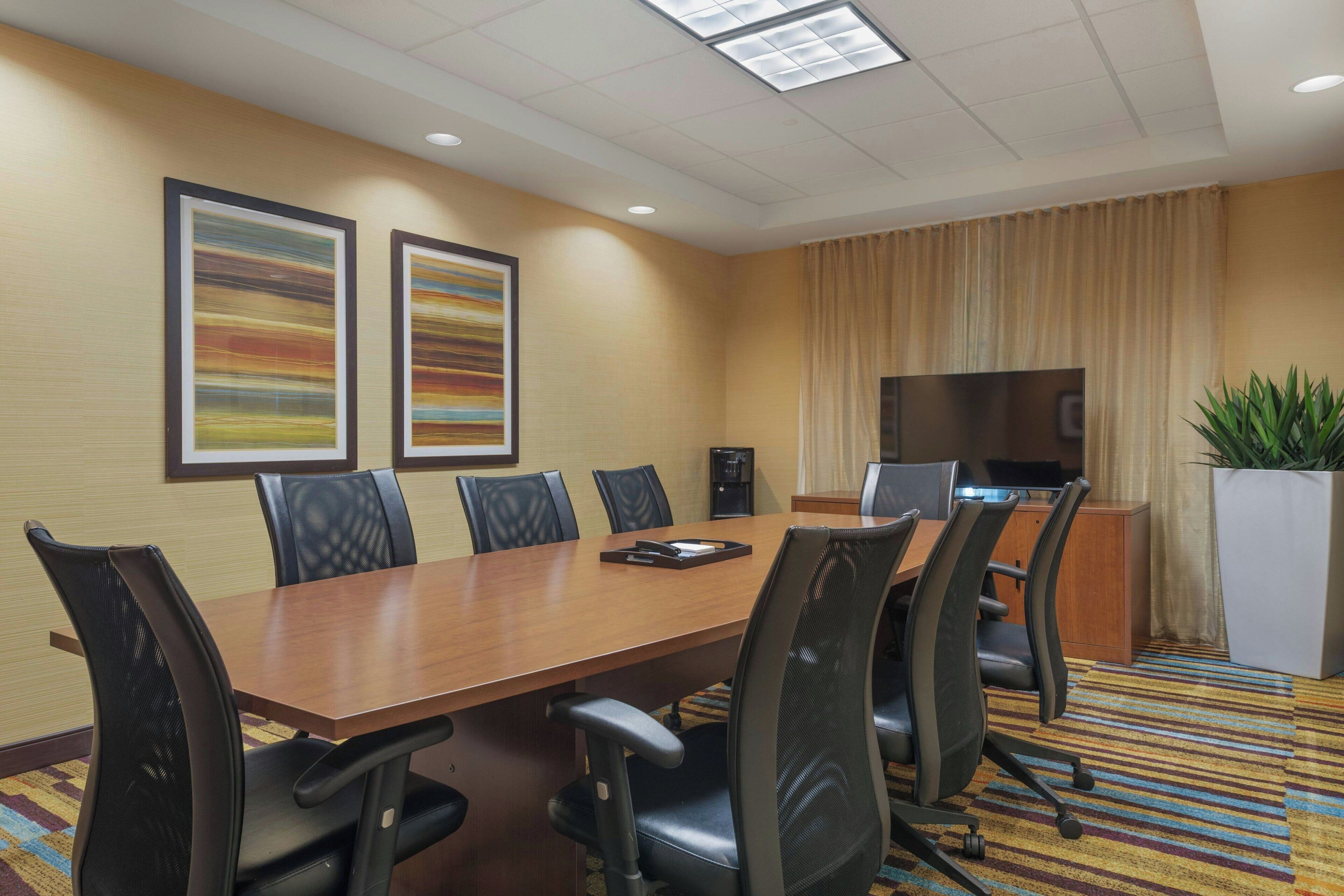 A conference room with a long table, six chairs, two framed artworks, a television, and a potted plant.