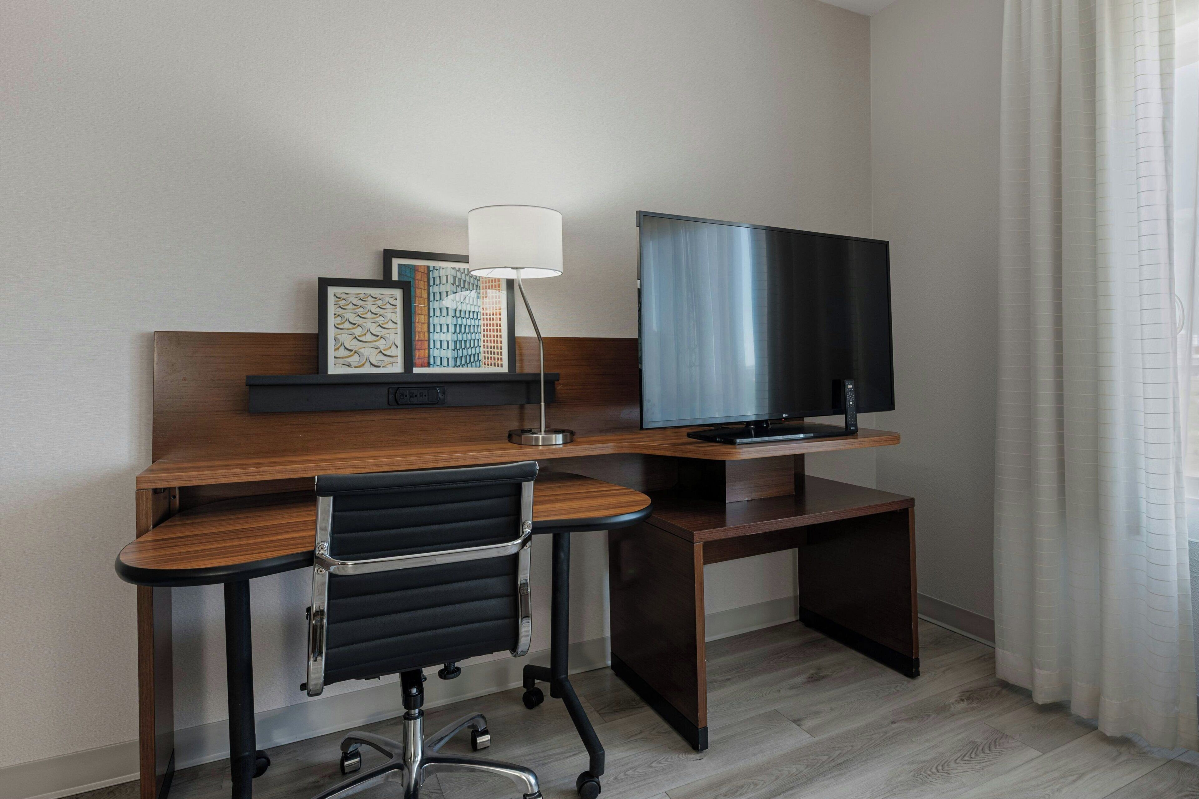 A hotel room with a wooden desk, a black office chair, a flat-screen TV, and a lamp.
