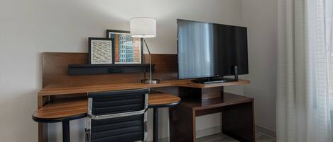 A hotel room with a wooden desk, a black office chair, a flat-screen TV, and a lamp.
