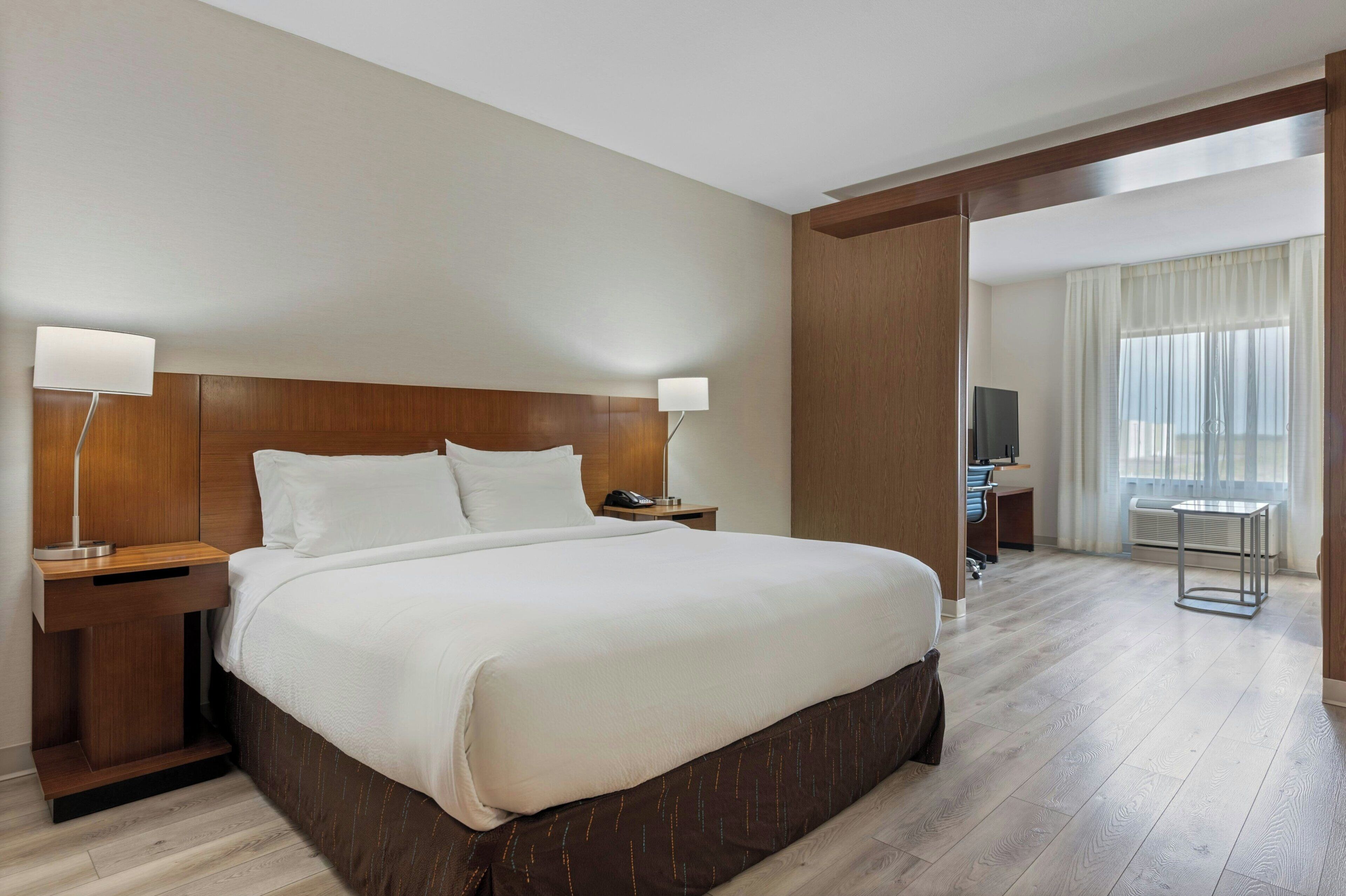 A hotel room with a large bed, wooden headboard, bedside lamps, a desk, and a view of a cityscape through the window.