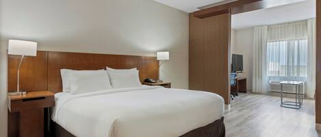A hotel room with a large bed, wooden headboard, bedside lamps, a desk, and a view of a cityscape through the window.