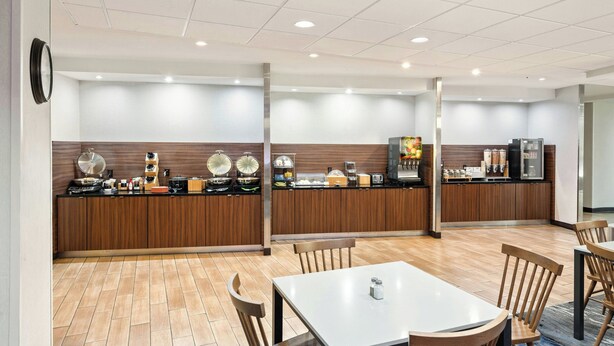 A modern hotel kitchen with wooden cabinets, a central island, and a dining area with wooden chairs and a table. - Fairfield Inn & Suites Alamosa (Alamosa)