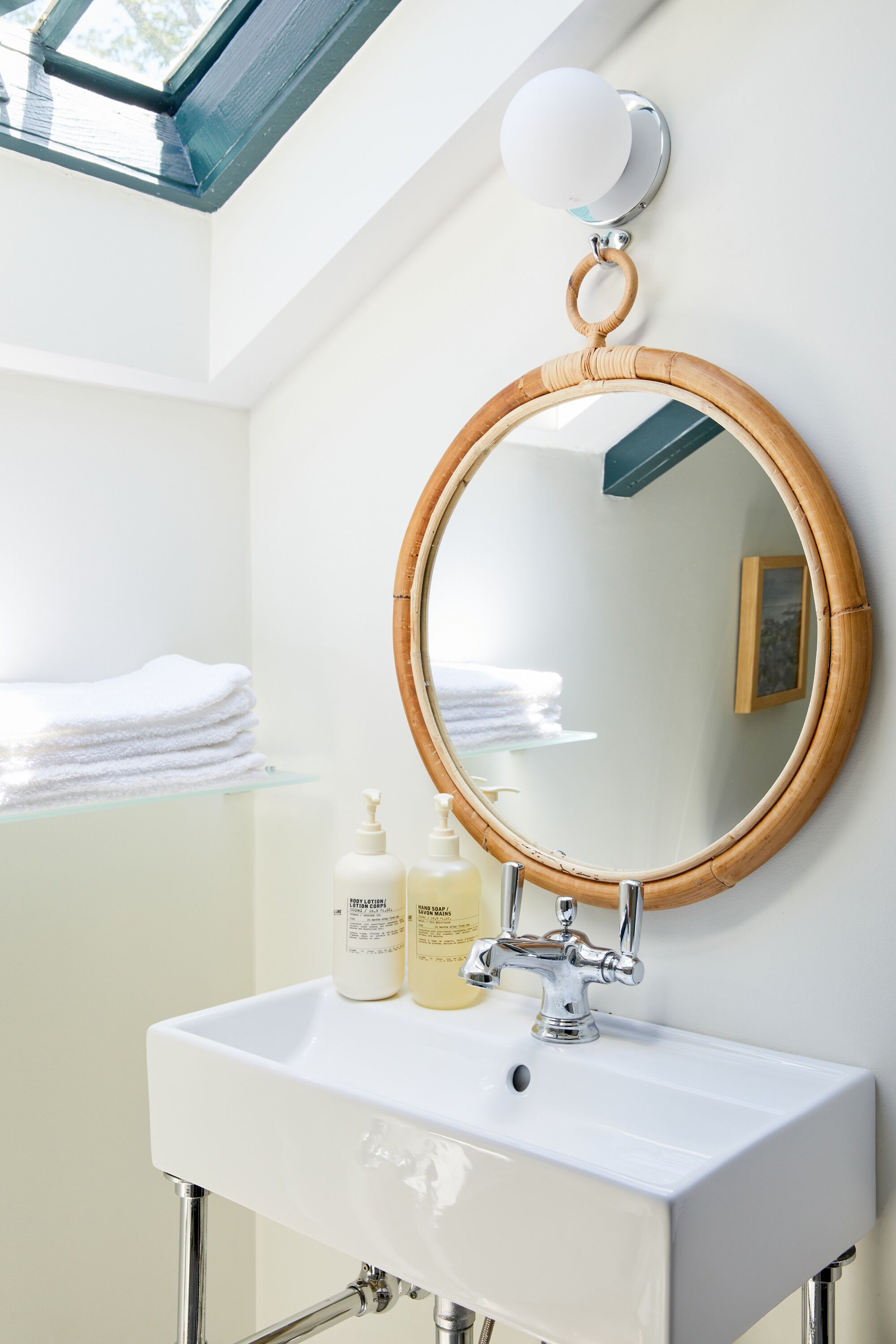 Une salle de bain avec un lavabo blanc, un miroir rond encadré de bambou et une étagère en verre où sont pliées des serviettes.