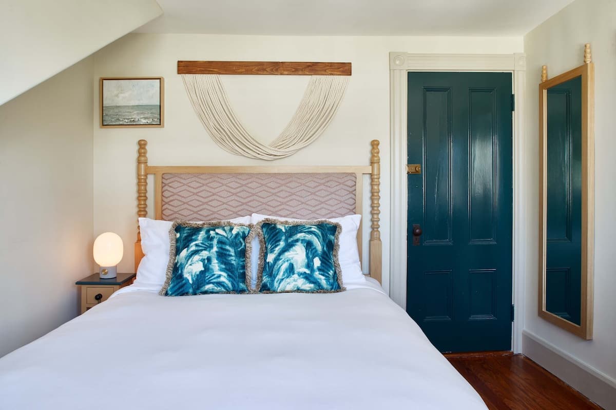 A bedroom with a bed, a nightstand with a lamp, a wooden headboard, a macramé wall hanging, a framed picture, and a dark door.