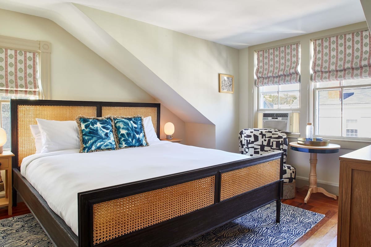 A bedroom with a bed, a bedside table, a lamp, a chair, a small table, and a window with curtains.