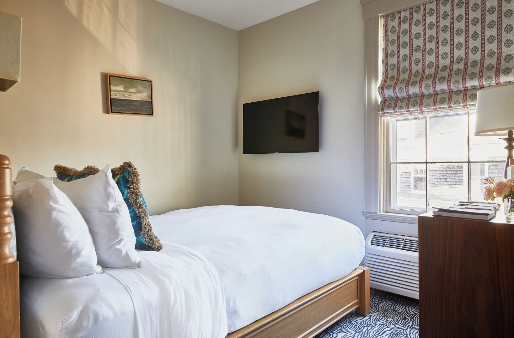Un dormitorio con cama, televisor de pantalla plana, ventana con cortinas y mesita de noche con lámpara y un libro.