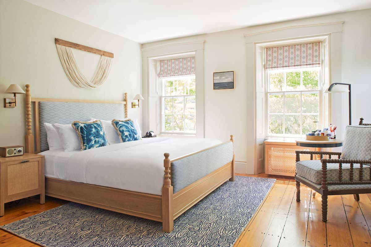 A bedroom with a wooden bed, a nightstand, a chair, and two windows with curtains.